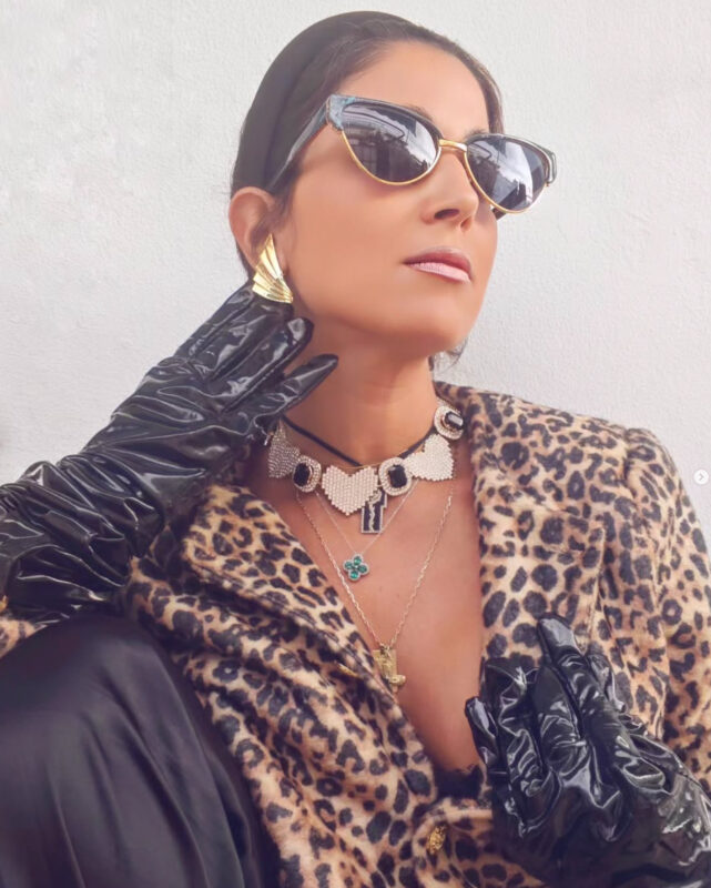 Γυναίκα με λεοπάρ παλτό, δερμάτινο παντελόνι, γάντια, τσόκερ και χρυσά σκουλαρίκια σε animal print και δερμάτινα look Double Trouble.