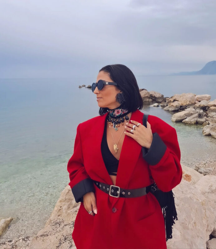 Ελένα με κόκκινο oversized παλτό, statement τσόκερ και κοσμήματα δίπλα στη θάλασσα, δείχνοντας πώς να μείνεις πιστή στο δικό σου στυλ μέσα στα trends.