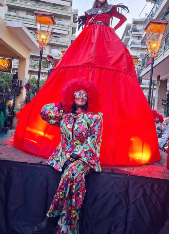 Πατρινό Καρναβάλι με πολύχρωμο κοστούμι, κόκκινη περούκα και statement γυαλιά μπροστά από τεράστια κόκκινη φούστα.