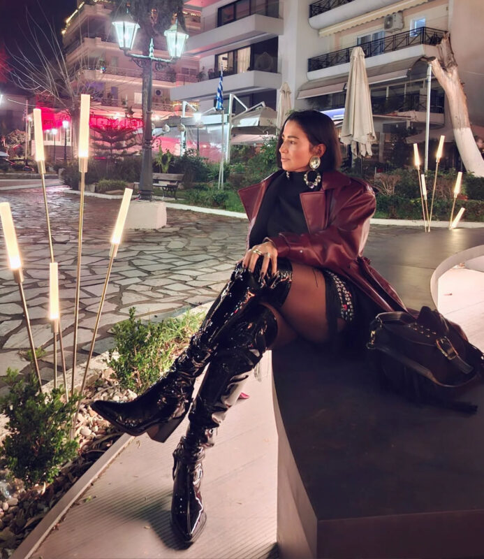 Γυναίκα με mini φόρεμα, over-the-knee λουστρίν μπότες, μπορντό παλτό και statement κοσμήματα σε βραδινό casual αλλά sexy look στην πόλη.