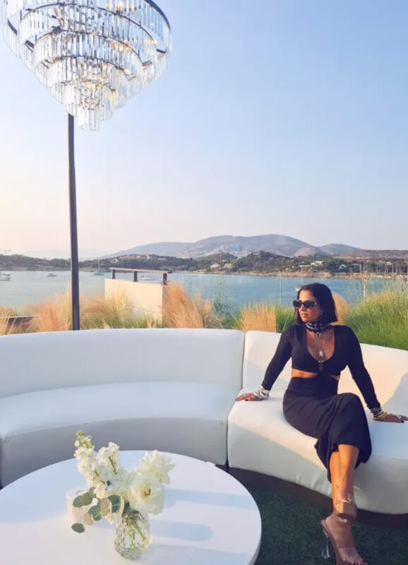 Γυναίκα με μαύρο φόρεμα και statement κολιέ κάθεται σε λευκό καναπέ στο Four Seasons με φόντο θάλασσα και πολυτελές φωτιστικό.