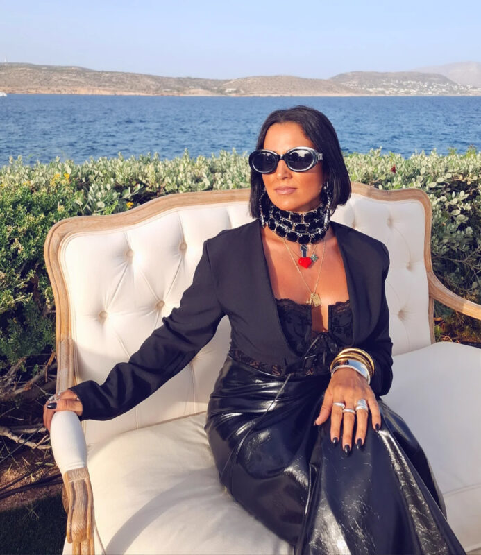 υναίκα με μαύρο δερμάτινο φόρεμα και statement τσόκερ κάθεται σε λευκό καναπέ μπροστά από τη θάλασσα στην Αθηναϊκή Ριβιέρα.