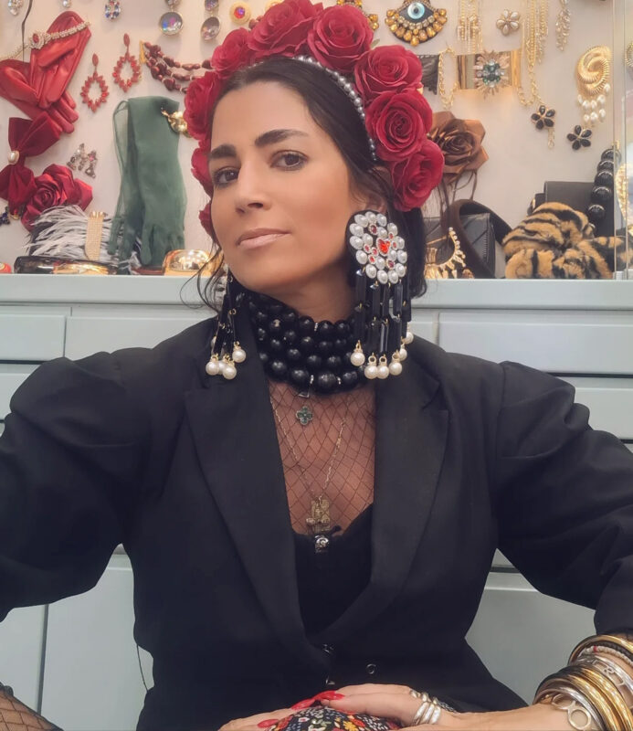 Γυναίκα με μαύρο σακάκι, στέκα με κόκκινα τριαντάφυλλα, μαύρο τσόκερ και μεγάλα κρεμαστά σκουλαρίκια με πέρλες μπροστά από βιτρίνα με κοσμήματα.