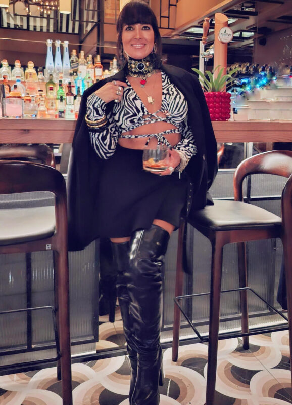 Γυναίκα με zebra crop top, μίνι φούστα, over the knee μπότες και statement τσόκερ σε cocktail bar με ποτό στο χέρι.