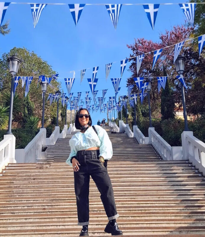 25 Μαρτίου με jeans, t-shirt και vintage σκουλαρίκια σε σκαλιά με ελληνικές σημαίες.