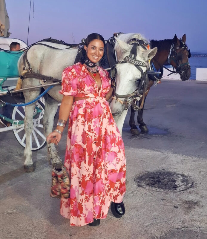 εικόνας: Γυναίκα στις Σπέτσες με ροζ φλοράλ φόρεμα, statement τσόκερ και βραχιόλια δίπλα σε άμαξα με άλογα στο ηλιοβασίλεμα