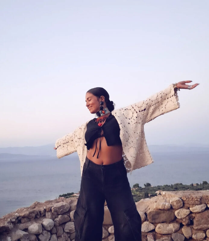 Γυναίκα σε θέα θάλασσας με μαύρο crop top, πλεκτό κιμονό και statement κολιέ, ανοίγει τα χέρια στον αέρα