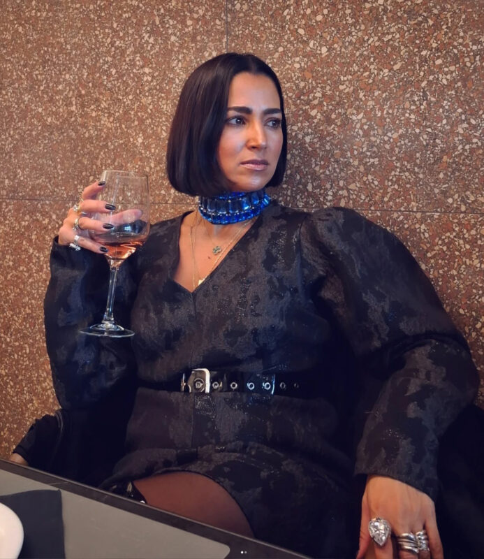 Γυναίκα σε εστιατόριο για δείπνο με μαύρο outfit, ζώνη και μπλε statement τσόκερ, κρατώντας ποτήρι κρασί