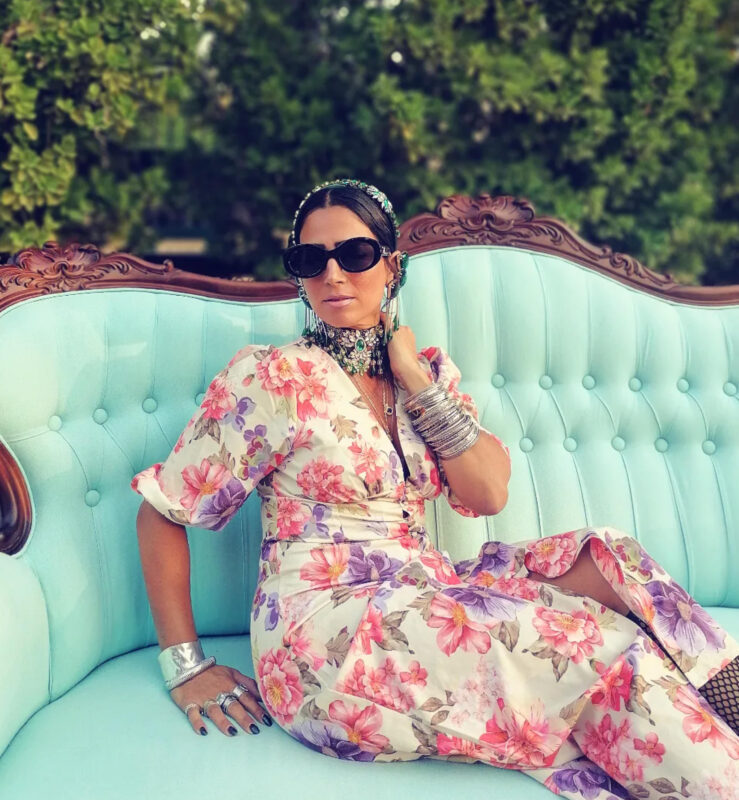 Έλενα με φλοράλ φόρεμα με κοσμήματα, statement τσόκερ, μεγάλα σκουλαρίκια και stacked βραχιόλια σε κήπο.