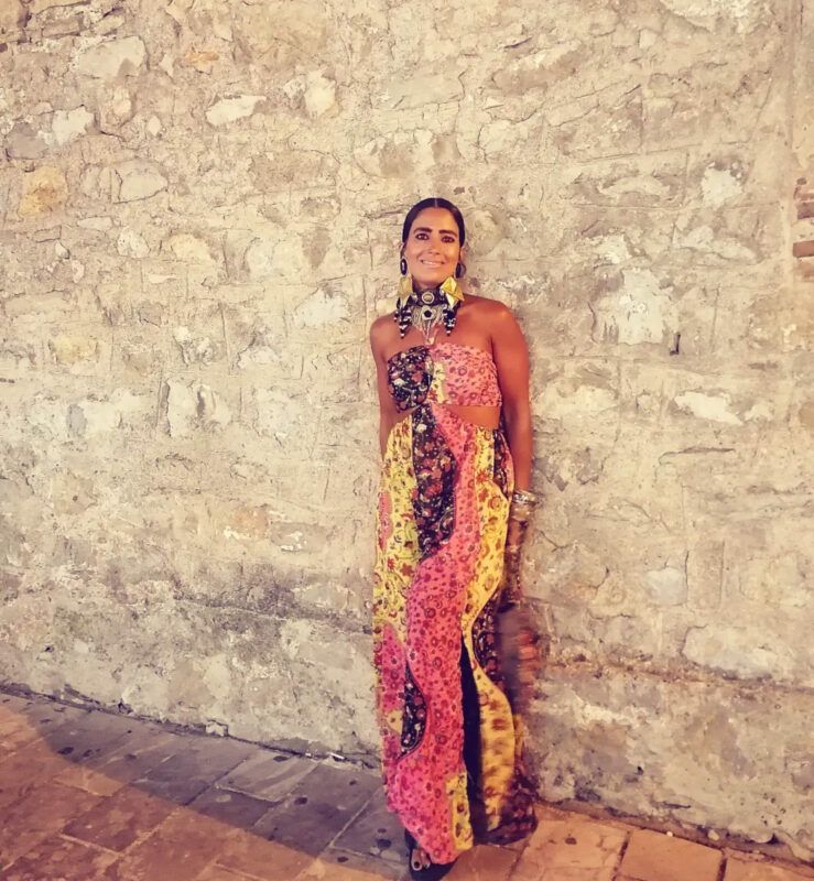 Πολύχρωμο καλοκαιρινό φόρεμα με τσόκερ, μεγάλα statement σκουλαρίκια και πολλά βραχιόλια μπροστά σε πέτρινο τοίχο.