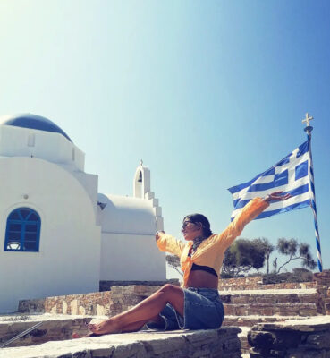 Διακοπές σε νησί με τσόκερ, denim φούστα και χρωματιστό top μπροστά σε εκκλησάκι και ελληνική σημαία.