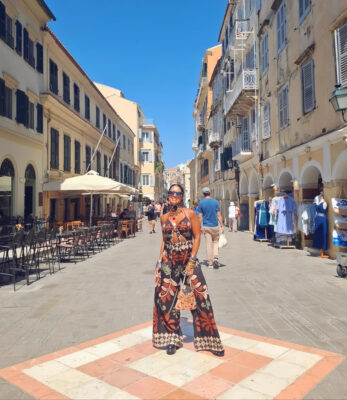 Γυναίκα σε διακοπές σε νησί με printed ολόσωμη φόρμα, statement τσόκερ, layered κολιέ, πολλά βραχιόλια και μικρό τσαντάκι.