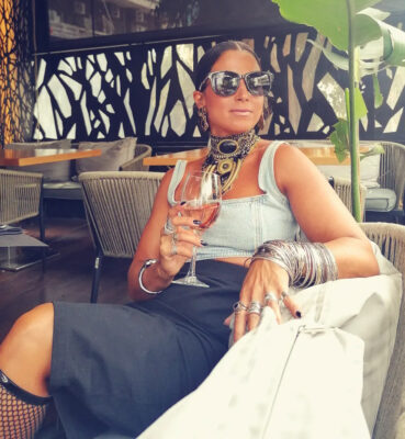 Γυναίκα σε aperitivo look με statement τσόκερ, denim top, μαύρη φούστα, πολλά ασημί βραχιόλια και ποτήρι κρασί.