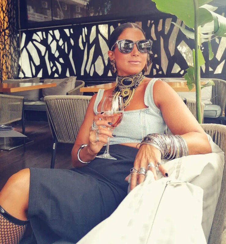 Γυναίκα σε aperitivo look με statement τσόκερ, denim top, μαύρη φούστα, πολλά ασημί βραχιόλια και ποτήρι κρασί.