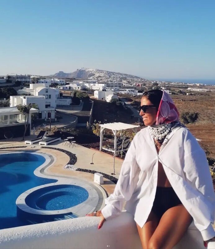 Γυναίκα στη Σαντορίνη με ροζ φουλάρι στα μαλλιά, λευκό oversized πουκάμισο, statement κολιέ με πολλές πέρλες και μαύρα γυαλιά ηλίου.