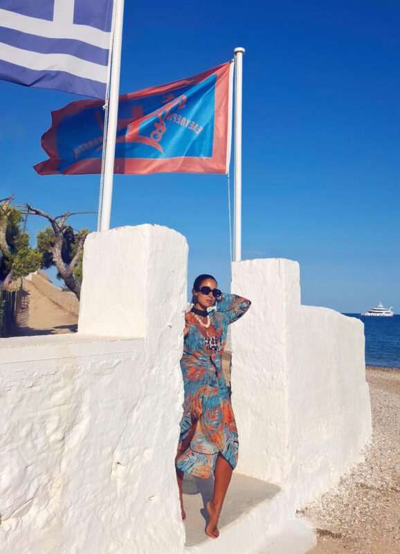 Γυναίκα στις Σπέτσες με πολύχρωμο boho φόρεμα πάνω από animal-print μαγιό, statement κολιέ, βραχιόλια και γυαλιά ηλίου δίπλα στη θάλασσα.
