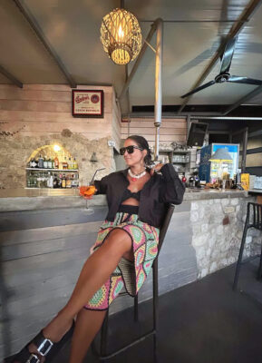 Γυναίκα σε καλοκαιρινό bar με μαύρο top, πολύχρωμο printed παντελόνι, layered κολιέ, πολλά βραχιόλια και cocktail στο χέρι.