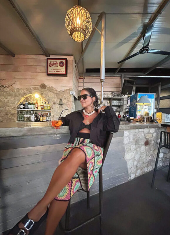 Γυναίκα σε καλοκαιρινό bar με μαύρο top, πολύχρωμο printed παντελόνι, layered κολιέ, πολλά βραχιόλια και cocktail στο χέρι.