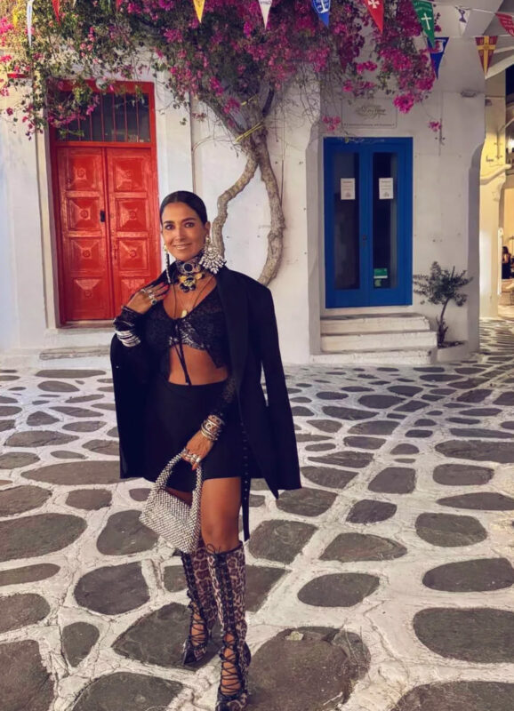 Γυναίκα σε σοκάκι της Μυκόνου με μαύρο outfit, statement τσόκερ, πολλά βραχιόλια, leopard ψηλές μπότες και μικρή μεταλλική τσάντα χειρός.