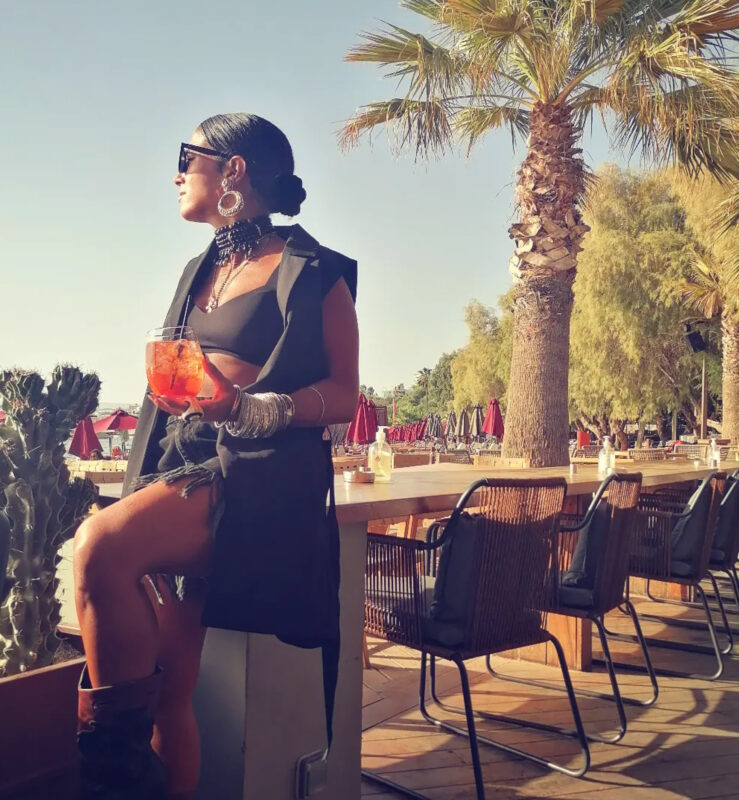 Γυναίκα σε sunset cocktail με total black look, αμάνικο blazer, statement choker, μεγάλα σκουλαρίκια, πολλά βραχιόλια και cocktail στο χέρι.
