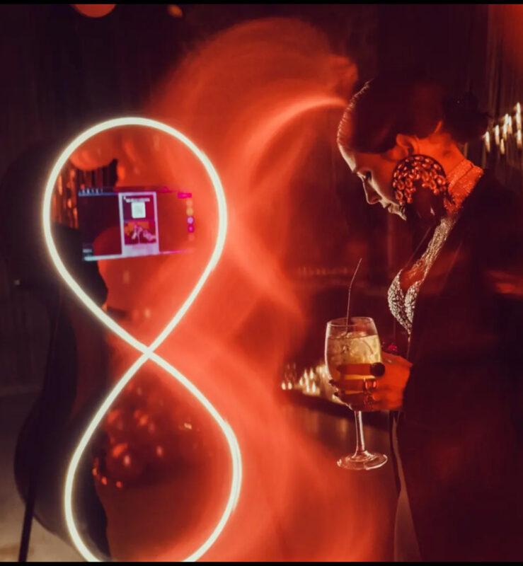 Γυναίκα ποζάρει μπροστά στο KIBELI photobooth με μαύρο βραδινό look, τεράστια statement σκουλαρίκια, μεταλλικό τσόκερ, layered κολιέ, δαχτυλίδια και cocktail στο χέρι.