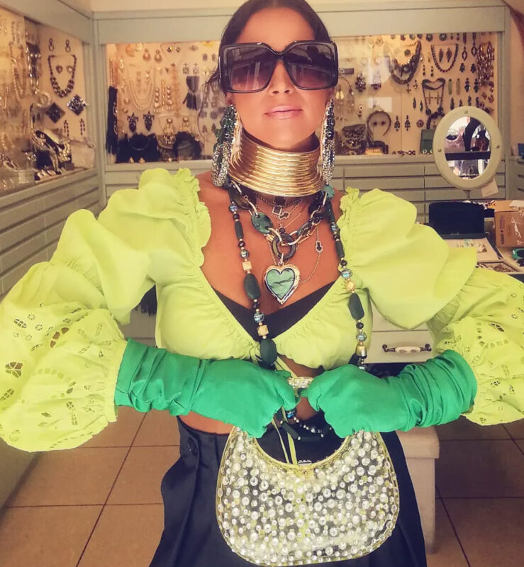 Γυναίκα μέσα στο κατάστημα Double Trouble στην Πάτρα με lime top με ογκώδη μανίκια, πράσινα μακριά γάντια, μεγάλο χρυσό τσόκερ, πράσινο κολιέ καρδιά, μεγάλα σκουλαρίκια και oversized μαύρα γυαλιά.