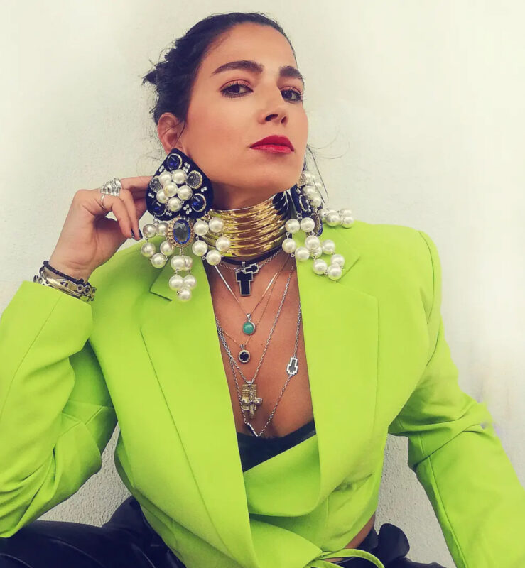 Γυναίκα με lime πράσινο blazer, πολύ μεγάλα σκουλαρίκια με πέρλες και μπλε πέτρες, φαρδύ χρυσό τσόκερ και πολλά layered κολιέ.