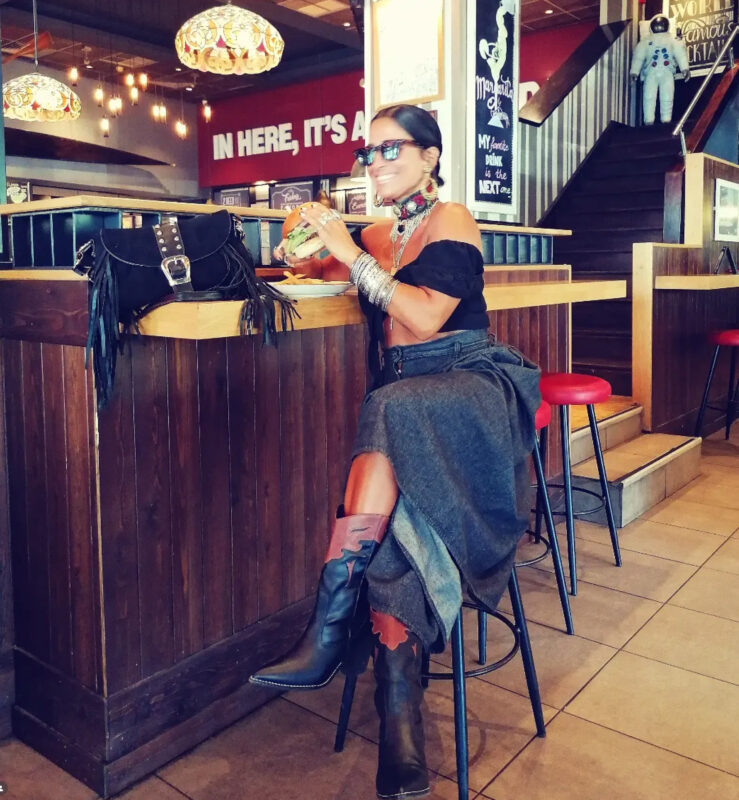 Γυναίκα σε bar με μακριά denim φούστα, μαύρο off-shoulder top, western μπότες, μεγάλα σκουλαρίκια, layered κολιέ και πολλά μεταλλικά βραχιόλια.