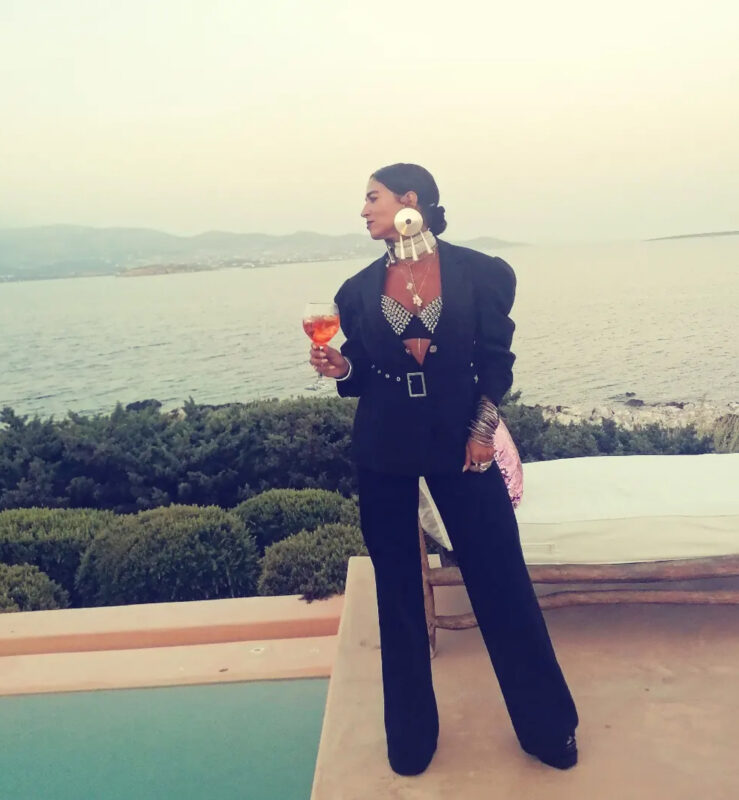 Γυναίκα σε παραθαλάσσιο resort με μαύρο oversized blazer, φαρδύ παντελόνι, λαμπερό bustier, XXL σκουλαρίκια, layered κολιέ και πολλά βραχιόλια.