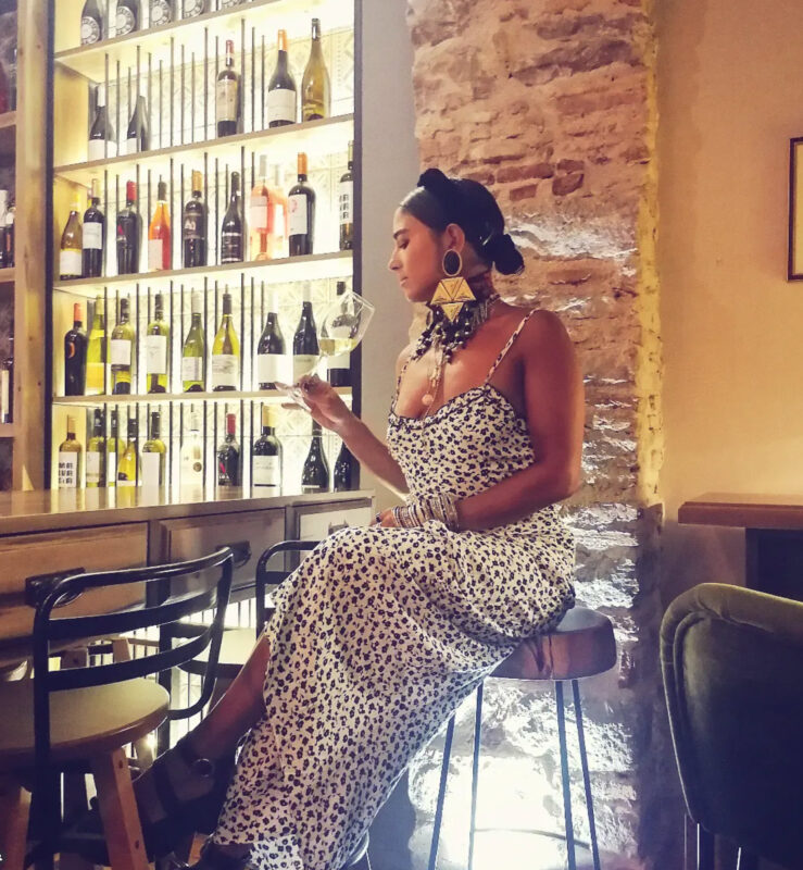 Γυναίκα σε wine bar με μακρύ printed φόρεμα, μεγάλα χρυσά γεωμετρικά σκουλαρίκια, τσόκερ, layered κολιέ και πολλά μεταλλικά βραχιόλια.