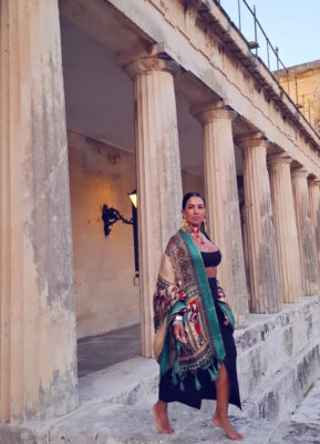 Γυναίκα στην Κέρκυρα με μαύρο outfit, μεγάλο πολύχρωμο printed φουλάρι στους ώμους, statement σκουλαρίκια, κολιέ και πολλά βραχιόλια.